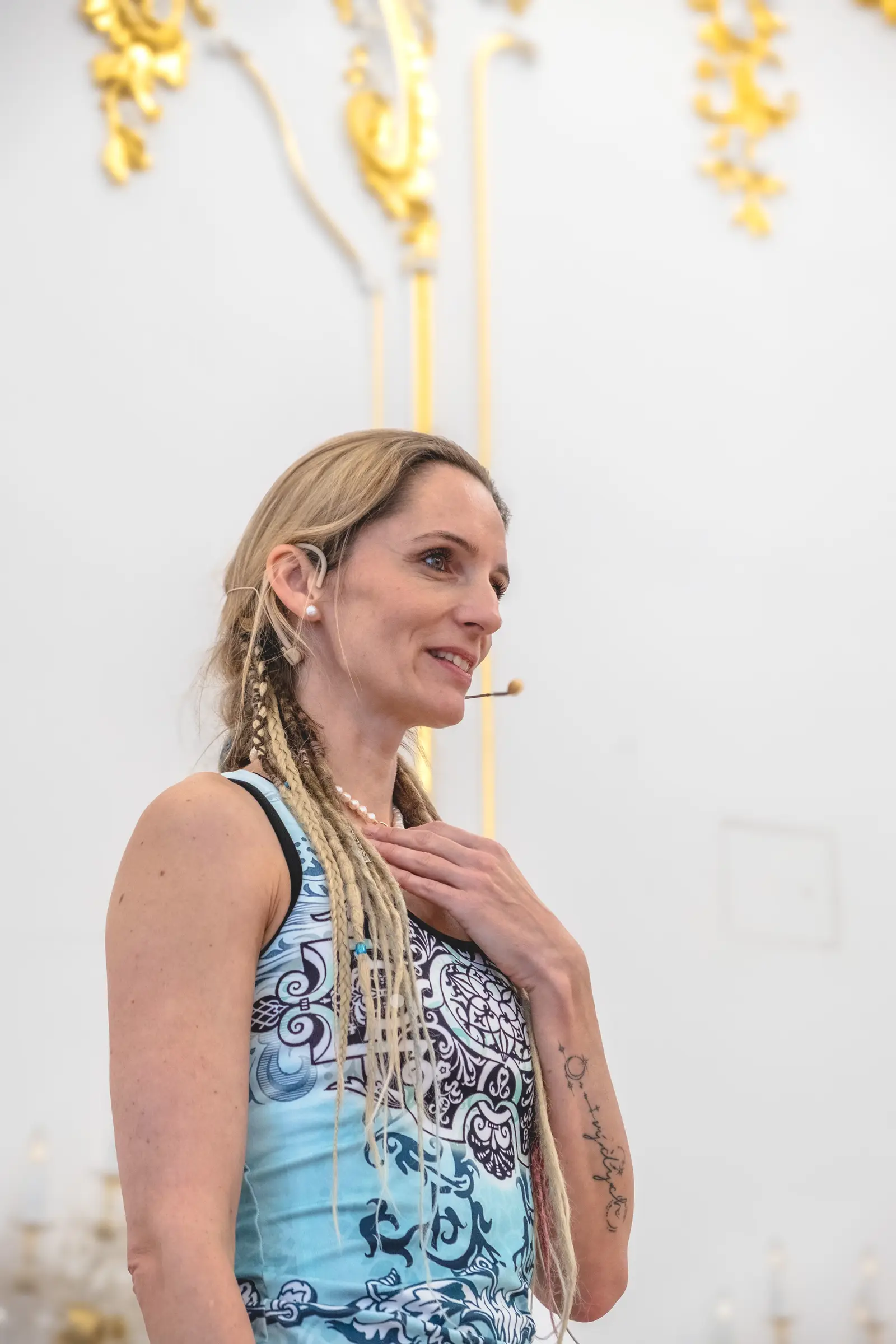 Žena s náhlavným mikrofónom a rukou na hrudi sa usmieva, za ňou biela stena so zlatou štukovou výzdobou.