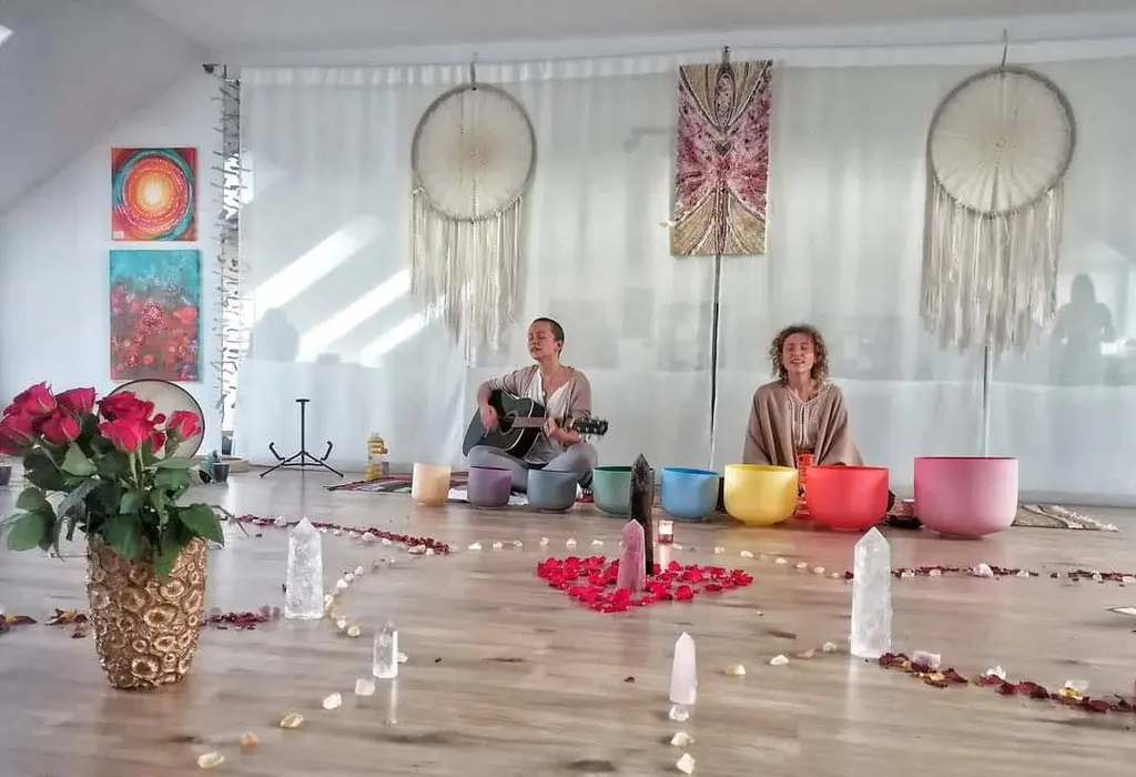 Dve ženy sedia na podlahe a spievajú, jedna hrá na gitare; pred nimi rad farebných kryštálových mís, kryštály a lupene ruží v tvare srdca, vľavo kytica ruží.